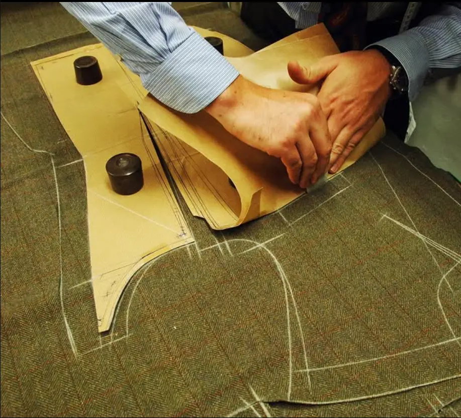 Image Credit: Knize Tailor’s hands working with a paper pattern on chalk-marked fabric.