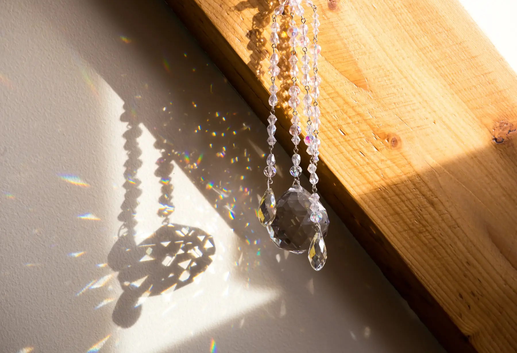 Crystal suncatcher casting rainbow reflections and shadows on a sunlit wall and a wooden beam.