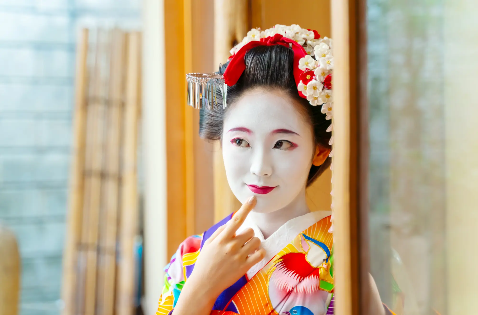 A Maiko, apprentice Geisha, in full white face makeup and traditional kimono, pointing to her cheek