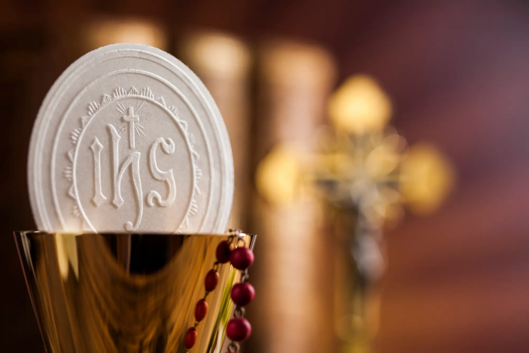 Eucharistic host engraved with the IHS monogram resting on a golden chalice, with a rosary and blurred cross in the background.